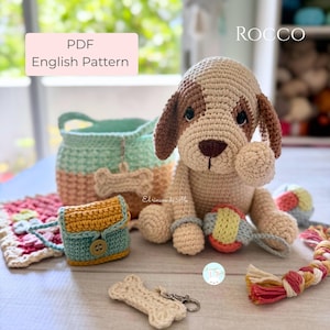 Puede incluir: Una colección de juguetes y accesorios para perros hechos a mano con ganchillo. Incluye un perro de peluche, una cesta, un pequeño bolso, un llavero con forma de hueso y un juguete de cuerda. El perro es beige y marrón. El texto "PDF English Pattern" es visible.