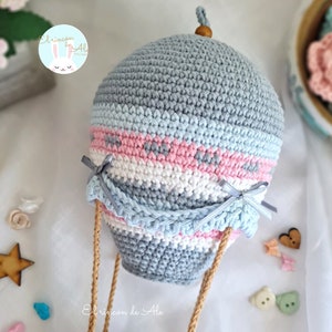 May include: A crocheted hot air balloon with a grey, blue, pink and white striped pattern. The balloon has a wooden top and is attached to brown strings.