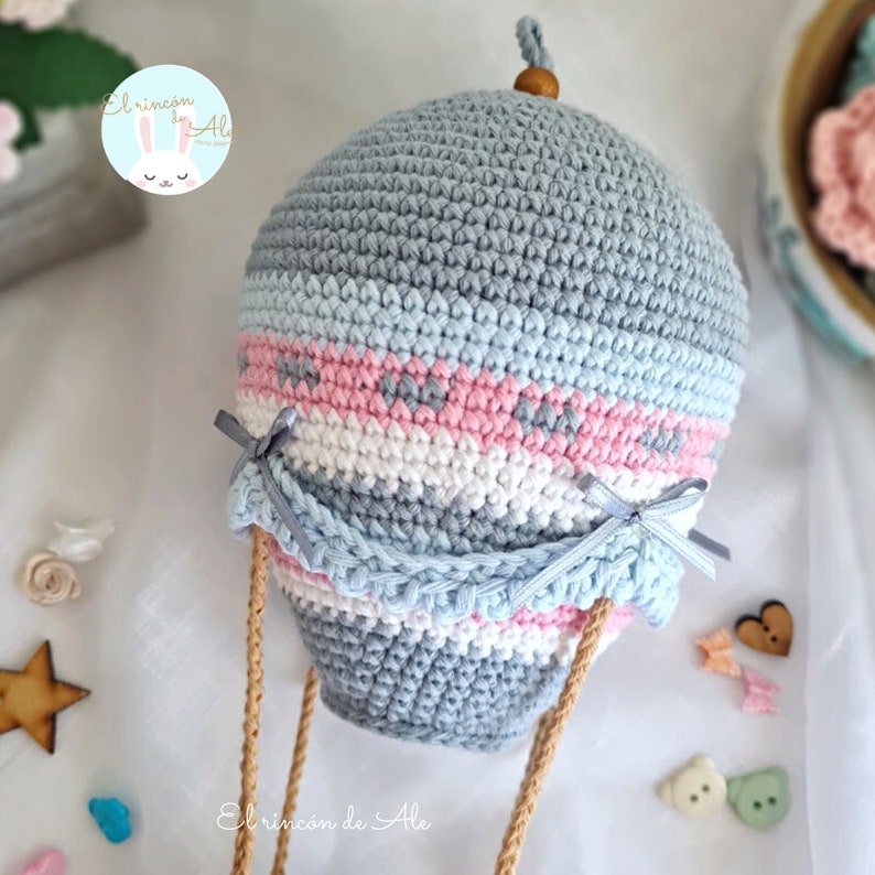 May include: A crocheted hot air balloon with a grey, blue, pink and white striped pattern. The balloon has a wooden top and is attached to brown strings.