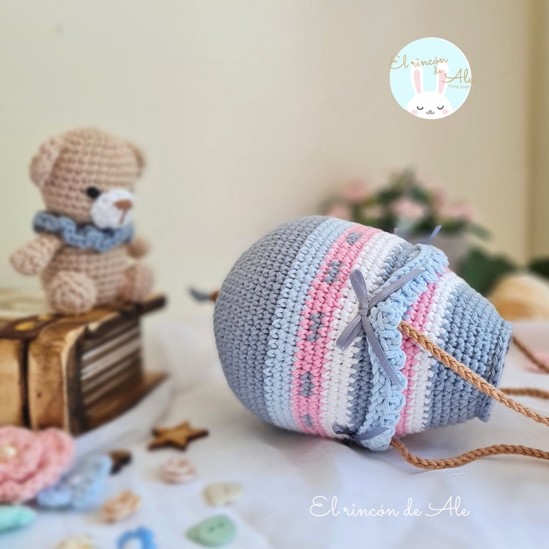 May include: A crocheted hot air balloon toy in shades of grey, blue, pink, and white with a brown rope handle. The balloon is lying on a white surface with a small brown teddy bear and other small crocheted items in the background.