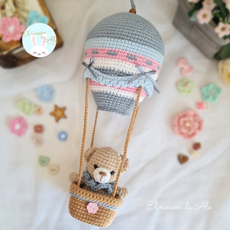 May include: A crocheted hot air balloon with a teddy bear in the basket. The balloon is grey, blue, pink, and white with a crocheted trim. The basket is brown with a pink flower.