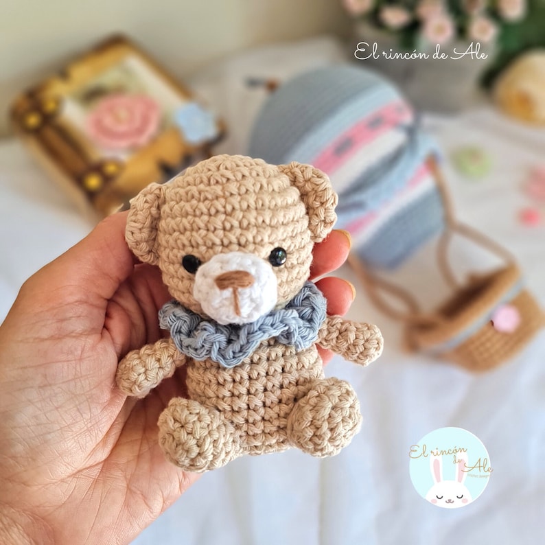May include: A crocheted teddy bear with a light brown body, white face, and blue scarf. The bear is sitting in a hand.