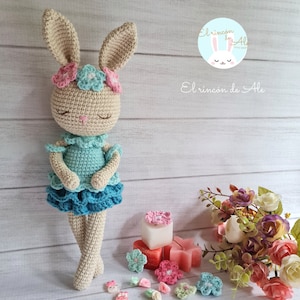 Puede incluir: Un conejito de crochet que lleva un vestido azul y verde con una corona de flores rosa y blanca. El conejito está de pie sobre una superficie de madera con un ramo de flores y pequeñas flores decorativas.