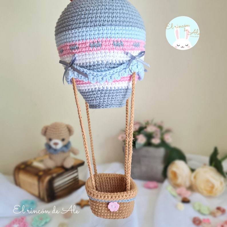 May include: A crocheted hot air balloon with a blue, pink and white striped balloon, a brown basket and a pink flower button. The balloon is hanging from a brown string.