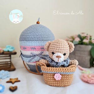 May include: A crocheted teddy bear wearing a blue scarf sits in a brown crocheted basket. The basket is attached to a grey and white striped hot air balloon with pink accents. The balloon has a brown wooden top.