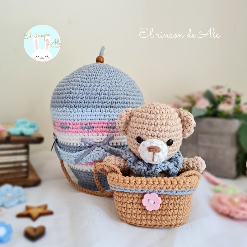 May include: A crocheted teddy bear wearing a blue scarf sits in a brown crocheted basket. The basket is attached to a grey and white striped hot air balloon with pink accents. The balloon has a brown wooden top.