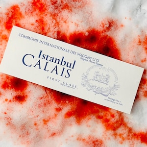 May include: A vintage "Istanbul Calais" first-class train ticket, printed in navy blue on a cream-coloured background. The ticket is set against a backdrop of white with vibrant red splatters, creating a striking contrast.