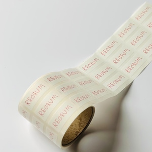 Puede incluir: Un rollo de etiquetas blanquecinas con la palabra "REGGUM" impresa en rojo. Las etiquetas son rectangulares y están parcialmente enrolladas, revelando el interior marrón del rollo. El fondo es blanco.