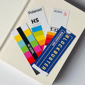 May include: A selection of vintage paper ephemera, including a Blockbuster membership card, Polaroid HS video cassette label, and a Sony T-12 label. They are arranged on a cream-coloured surface.