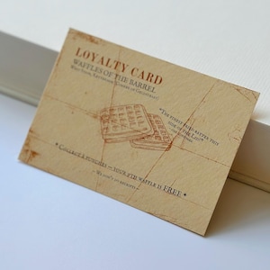 Peut inclure: Une carte de fidélité beige pour "Waffles of the Barrel" avec des illustrations de gaufres et du texte. La carte indique "Collectez 8 tampons - votre 9ème gaufre est GRATUITE". La carte est inclinée sur une surface blanche.