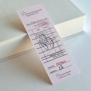 May include: A Bloomingdale's receipt with handwritten details, including "PAID CASH" and item descriptions like "Black Patent Leather Gloves." The receipt is pink with black text and rests on a white book.