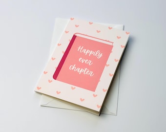 Pode incluir: Um cartão com um design de livro rosa e as palavras "Happily ever chapter" em escrita branca. O cartão é rodeado por uma borda de pequenos corações rosa e repousa sobre um envelope branco.