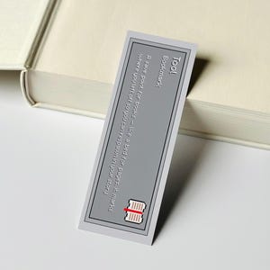 May include: A grey bookmark with a black border and text that reads "Tool Bookmark: A save point for books - like a bed for pages, it marks where you left off so you can respawn in your story." The bookmark is next to a book.