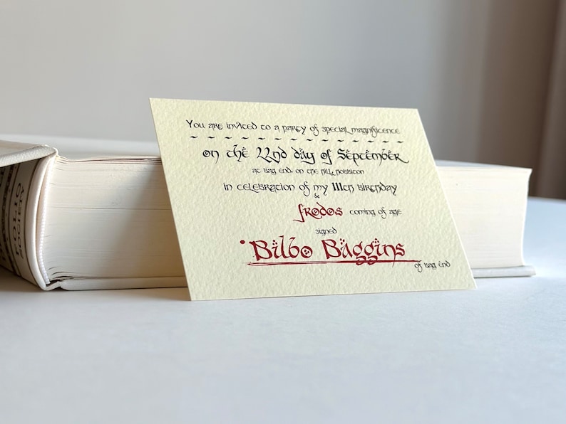 May include: A cream-colored invitation card with black calligraphy text. The text reads: "You are invited to a party of special magnificence on the 22nd day of September at Bag End on the Hill, Hobbiton in celebration of my 111th birthday & Frodo's coming of age. Signed Bilbo Baggins of Bag End."