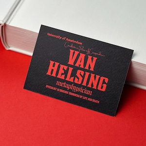 Könnte beinhalten: Eine schwarze Visitenkarte mit rotem Text "VAN HELSING, Metaphysiker, Spezialist für obskure Störungen des Lebens und des Todes". Die Karte liegt auf rotem Untergrund neben einem weißen Buch.
