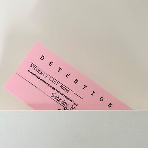 以下が含まれることがあります： ピンク色の留置票で、上部に「DETENTION」と、その下に「STUDENT'S LAST NAME」と書いてあります。この票には、生徒の姓と留置日の記入欄があり、日付欄には「Saturday, May」と記入されています。