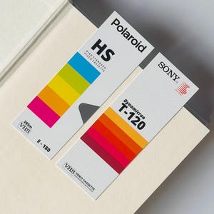 May include: Two vintage VHS video cassette tapes. One is labeled "Polaroid HS" with a rainbow color gradient, and the other is "SONY Dynamicron T-120" with a red to orange gradient. Both are on a light beige surface.