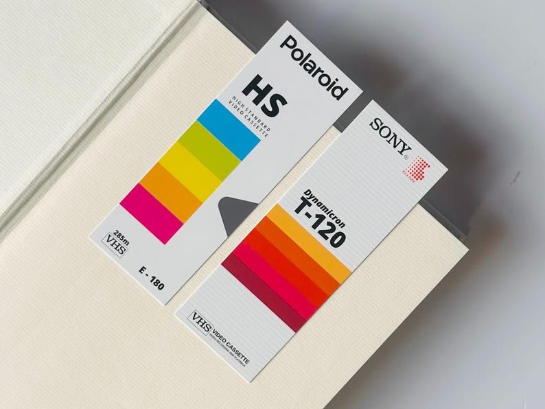 May include: Two vintage VHS video cassette tapes. One is labeled "Polaroid HS" with a rainbow color gradient, and the other is "SONY Dynamicron T-120" with a red to orange gradient. Both are on a light beige surface.