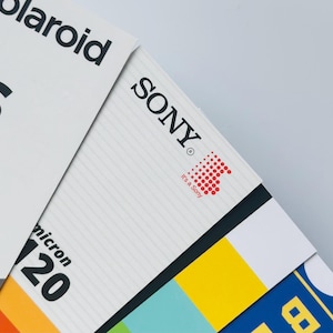 May include: Close-up of VHS and Sony cassette packaging. The VHS packaging is white with black text, including "VHS" and "Standard Cassette." The Sony packaging is white with the Sony logo and red dot design.