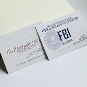 K&ouml;nnte beinhalten: Zwei Visitenkarten sind abgebildet, eine f&uuml;r Dr. Hannibal Lecter, Forensischer Psychiater, und die andere f&uuml;r das FBI. Die FBI-Karte zeigt das Siegel der Agentur und den Namen Will Graham. Beide Karten liegen auf einer hellen Oberfl&auml;che.