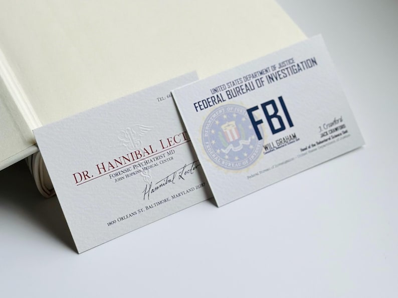 K&ouml;nnte beinhalten: Zwei Visitenkarten sind abgebildet, eine f&uuml;r Dr. Hannibal Lecter, Forensischer Psychiater, und die andere f&uuml;r das FBI. Die FBI-Karte zeigt das Siegel der Agentur und den Namen Will Graham. Beide Karten liegen auf einer hellen Oberfl&auml;che.