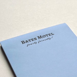 Op de afbeelding: Een lichtblauw notitieblok met de tekst "Bates Motel family friendly!" gedrukt op de voorkant.