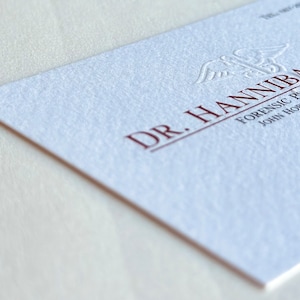 K&ouml;nnte beinhalten: Eine wei&szlig;e Visitenkarte mit gepr&auml;gtem Text in Dunkelrot. Die Karte tr&auml;gt die Aufschrift "DR. HANNIBAL LECTER" und enth&auml;lt den Titel "FORENSIC PSYCHIATRIST" und "JOHN HOPKINS MEDICAL". Die Karte hat eine strukturierte Oberfl&auml;che.