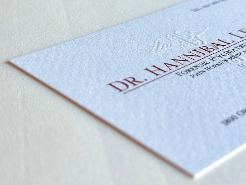 K&ouml;nnte beinhalten: Eine wei&szlig;e Visitenkarte mit gepr&auml;gtem Text in Dunkelrot. Die Karte tr&auml;gt die Aufschrift "DR. HANNIBAL LECTER" und enth&auml;lt den Titel "FORENSIC PSYCHIATRIST" und "JOHN HOPKINS MEDICAL". Die Karte hat eine strukturierte Oberfl&auml;che.