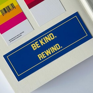 May include: A blue rectangular sticker with the text "BE KIND. REWIND." in yellow. The sticker has a gold border and is on a white surface. Other colorful rectangular cards are also visible.