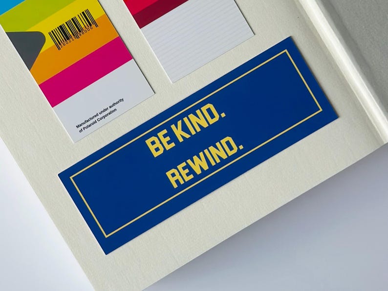 May include: A blue rectangular sticker with the text "BE KIND. REWIND." in yellow. The sticker has a gold border and is on a white surface. Other colorful rectangular cards are also visible.
