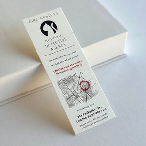 May include: A white bookmark with black text and a black cat silhouette. The text reads "Dirk Gently's Holistic Detective Agency". The bookmark also includes a map with a red circle marking a location. The text below the map reads "Somewhere here 33a Peckender St., London N1 01-359 9112 Phone today for the whole solution to your problem."
