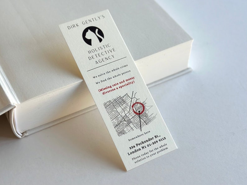 Op de afbeelding: Een witte bladwijzer met zwarte tekst en een zwart katten silhouet. De tekst luidt "Dirk Gently's Holistic Detective Agency". De bladwijzer bevat ook een kaart met een rode cirkel die een locatie markeert. De tekst onder de kaart luidt "Somewhere here 33a Peckender St., London N1 01-359 9112 Phone today for the whole solution to your problem."