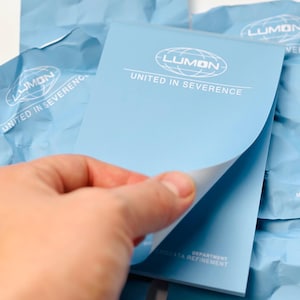 Op de afbeelding: Een lichtblauwe notitieblok met de tekst "LUMON UNITED IN SEVERENCE" in het wit. De notitieblok wordt vastgehouden, waarbij de bovenste pagina gedeeltelijk is opgetild. Andere notitieblokken en verpakkingen zijn zichtbaar op de achtergrond.