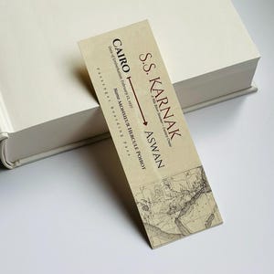 K&ouml;nnte beinhalten: Ein Lesezeichen im Vintage-Stil mit Text und Kartendesign, das auf einem cremefarbenen Buch liegt. Das Lesezeichen tr&auml;gt die Aufschrift "S.S. KARNAK" mit den Zielen "CAIRO" und "ASWAN" sowie den Namen "MONSIEUR HERCULE POIROT".