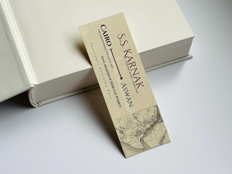 K&ouml;nnte beinhalten: Ein Lesezeichen im Vintage-Stil mit Text und Kartendesign, das auf einem cremefarbenen Buch liegt. Das Lesezeichen tr&auml;gt die Aufschrift "S.S. KARNAK" mit den Zielen "CAIRO" und "ASWAN" sowie den Namen "MONSIEUR HERCULE POIROT".