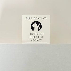 Op de afbeelding: Een witte kaart met zwarte tekst en een zwart silhouet van een kat. De tekst luidt "Dirk Gently's Holistic Detective Agency."