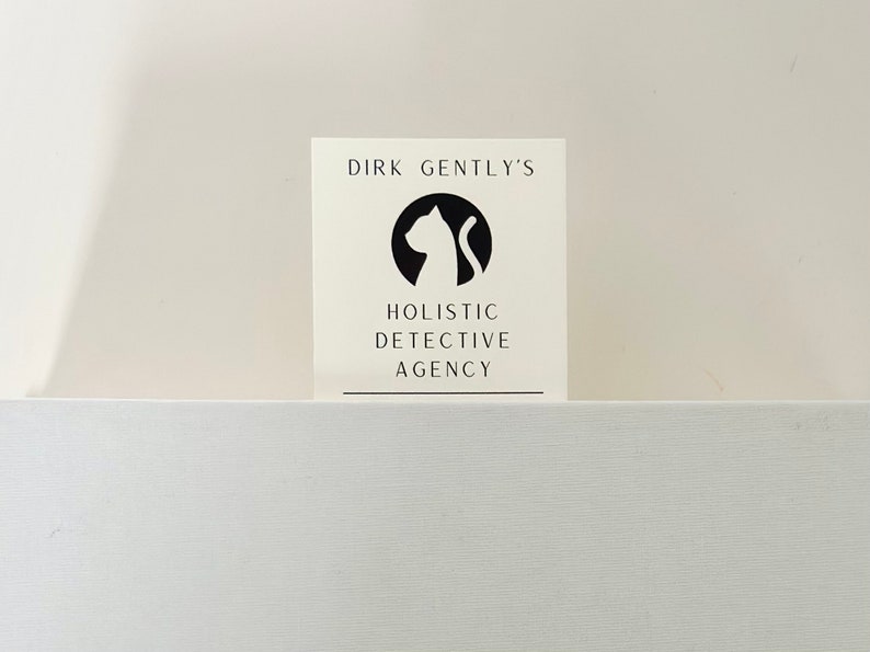 Op de afbeelding: Een witte kaart met zwarte tekst en een zwart silhouet van een kat. De tekst luidt "Dirk Gently's Holistic Detective Agency."