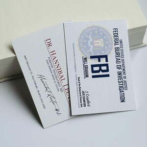 K&ouml;nnte beinhalten: Zwei Visitenkarten mit Text, eine f&uuml;r Dr. Hannibal Lecter, Forensischer Psychiater, und die andere f&uuml;r Will Graham, FBI. Die FBI-Karte zeigt das Siegel des US-Justizministeriums. Beide Karten liegen auf einer wei&szlig;en Oberfl&auml;che.