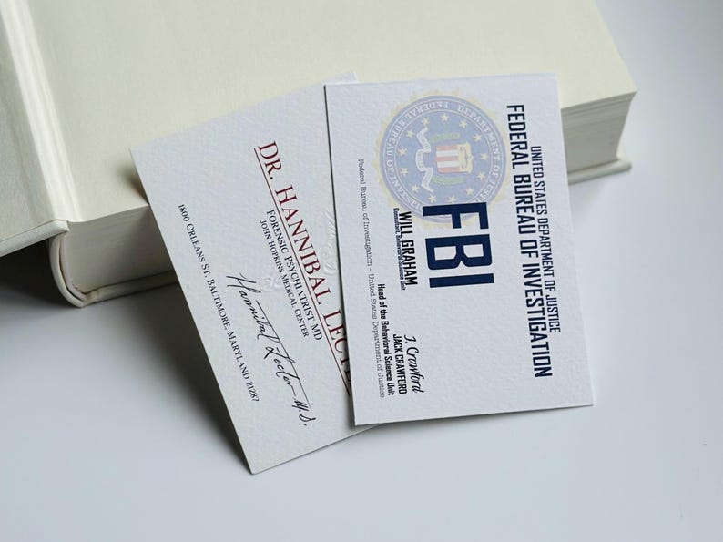 K&ouml;nnte beinhalten: Zwei Visitenkarten mit Text, eine f&uuml;r Dr. Hannibal Lecter, Forensischer Psychiater, und die andere f&uuml;r Will Graham, FBI. Die FBI-Karte zeigt das Siegel des US-Justizministeriums. Beide Karten liegen auf einer wei&szlig;en Oberfl&auml;che.