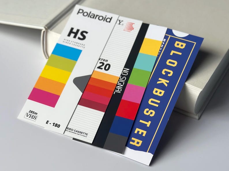 May include: A stack of retro-themed cards featuring VHS tape designs. The cards display the words "Polaroid", "HS", "cron 20", "NO SIGNAL", and "BLOCKBUSTER". The color palette includes vibrant hues of yellow, orange, pink, green, and blue.