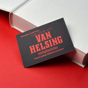 K&ouml;nnte beinhalten: Eine schwarze Visitenkarte mit rotem Text "VAN HELSING, Metaphysiker, Spezialist f&uuml;r obskure St&ouml;rungen des Lebens und des Todes". Die Karte liegt auf einer roten Oberfl&auml;che neben einem wei&szlig;en Buch.