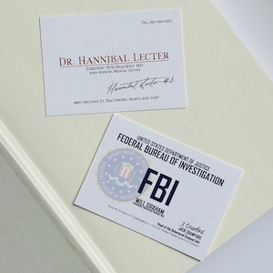K&ouml;nnte beinhalten: Zwei Visitenkarten auf einer cremefarbenen Oberfl&auml;che. Eine Karte lautet "Dr. Hannibal Lecter, Forensischer Psychiater MD" mit Adresse und Unterschrift. Die andere Karte ist eine FBI-Identifikationskarte mit dem Namen "Will Graham".