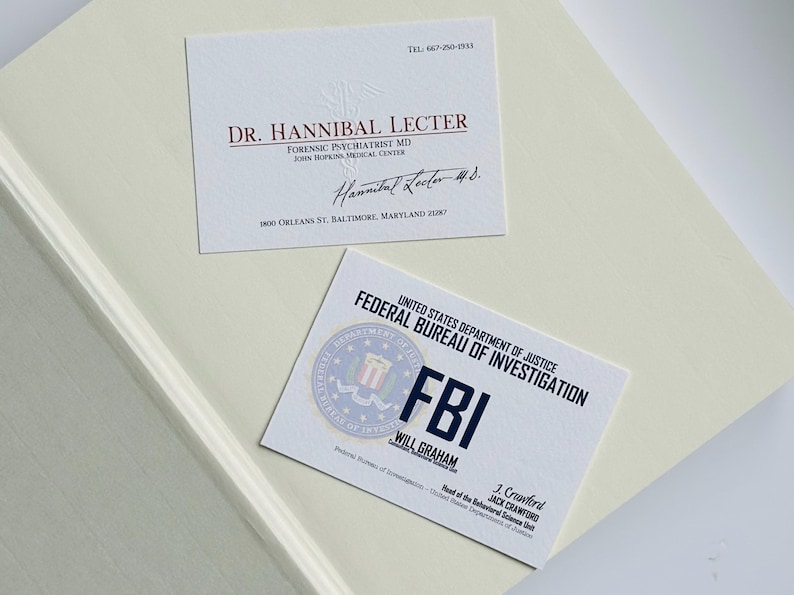 K&ouml;nnte beinhalten: Zwei Visitenkarten auf einer cremefarbenen Oberfl&auml;che. Eine Karte lautet "Dr. Hannibal Lecter, Forensischer Psychiater MD" mit Adresse und Unterschrift. Die andere Karte ist eine FBI-Identifikationskarte mit dem Namen "Will Graham".