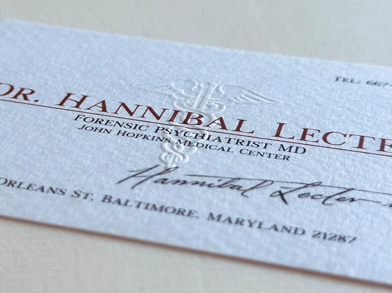 K&ouml;nnte beinhalten: Eine Visitenkarte mit dem Namen "Dr. Hannibal Lecter" in dunkelroter Schrift. Die Karte enth&auml;lt die Berufsbezeichnung "Forensischer Psychiater MD" und die Adresse des John Hopkins Medical Center in Baltimore, Maryland. Die Karte hat einen strukturierten, cremefarbenen Hintergrund.