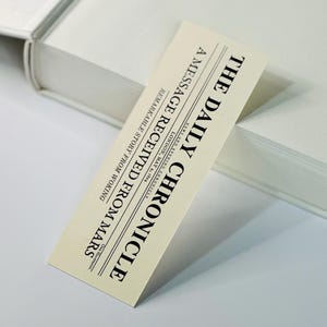 May include: A vintage-style bookmark in cream, featuring the headline "THE DAILY CHRONICLE" in black. The text "A MESSAGE RECEIVED FROM MARS" and "REMARKABLE STORY FROM WOKING" are also visible. The bookmark rests on a white book.