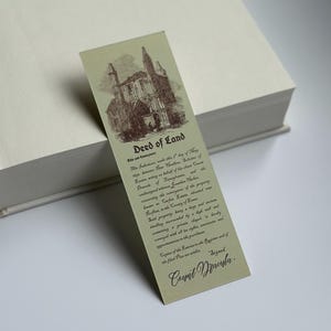 May include: A vintage-style bookmark with a sepia-toned illustration of a building and text. The bookmark reads "Deed of Land" and includes additional text, with the signature "Count Dracula" at the bottom. The bookmark is resting on a white book.