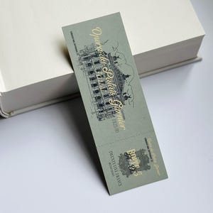 Puede incluir: Un marcapáginas verde salvia con letras doradas y una ilustración detallada de un edificio. El marcapáginas está apoyado en un libro blanco. El texto del marcapáginas dice "Opéra du Palais Garnier".