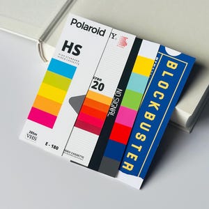 May include: A retro-style VHS video cassette label with the Polaroid and Blockbuster logos. The label features a colorful spectrum, the text "HS", "cron 20", "NO SIGNAL", and "E-180". The design evokes nostalgia for the 1980s and 1990s.