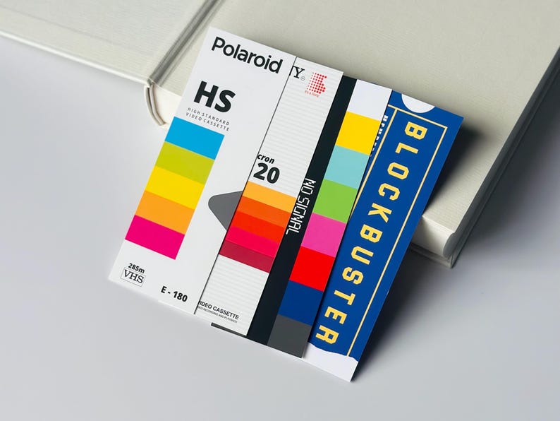 May include: A retro-style VHS video cassette label with the Polaroid and Blockbuster logos. The label features a colorful spectrum, the text "HS", "cron 20", "NO SIGNAL", and "E-180". The design evokes nostalgia for the 1980s and 1990s.