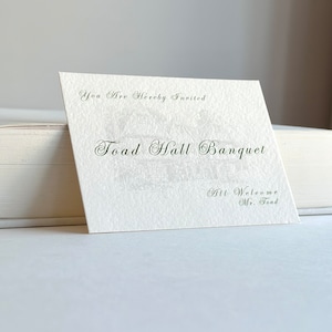 Puede incluir: Una tarjeta blanca con texto negro que dice "You Are Hereby Invited" y "Toad Hall Banquet". La tarjeta también tiene el texto "All Welcome Mr. Toad" en una fuente más pequeña.
